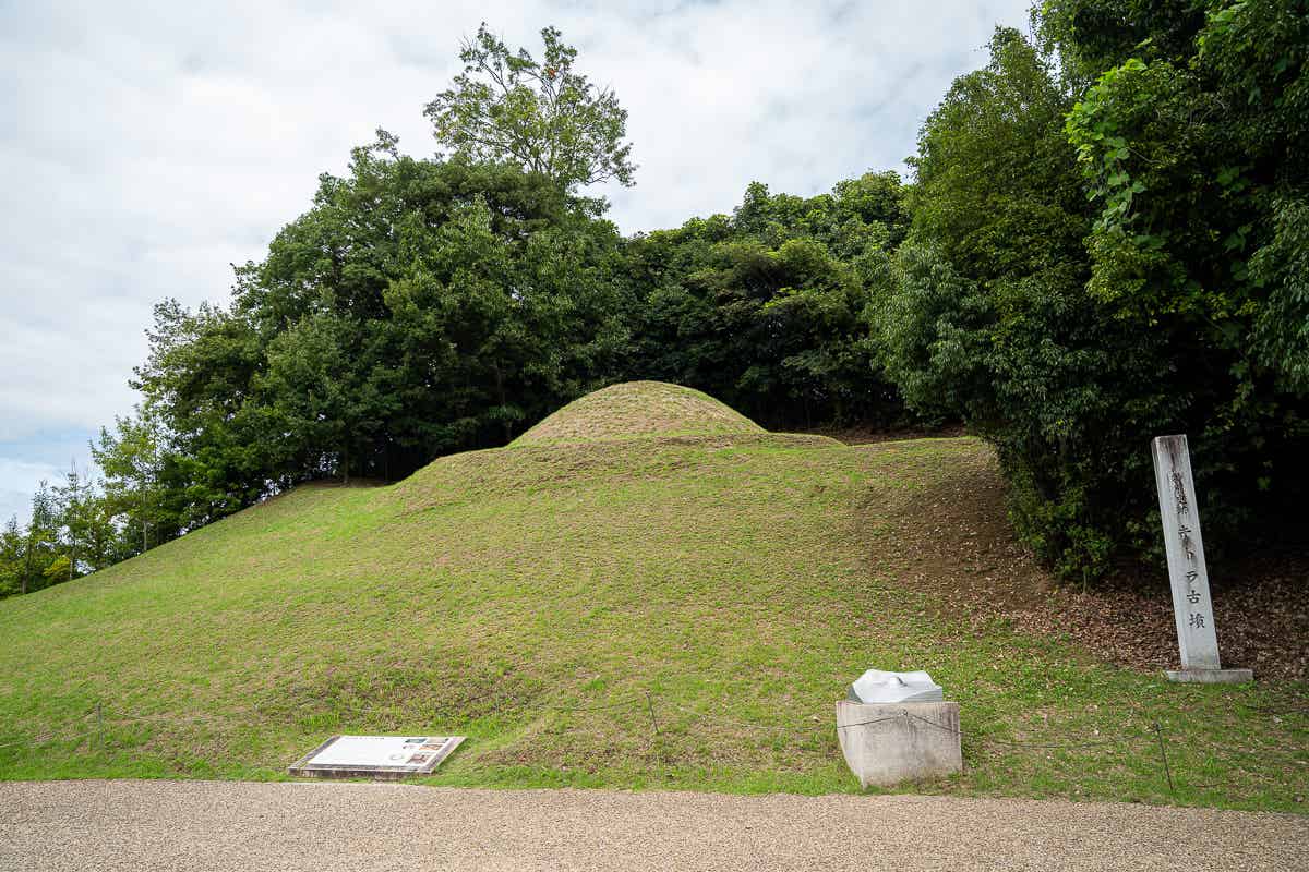 木々に囲まれた芝生の丸い古墳と前に立つ石碑（キトラ古墳）