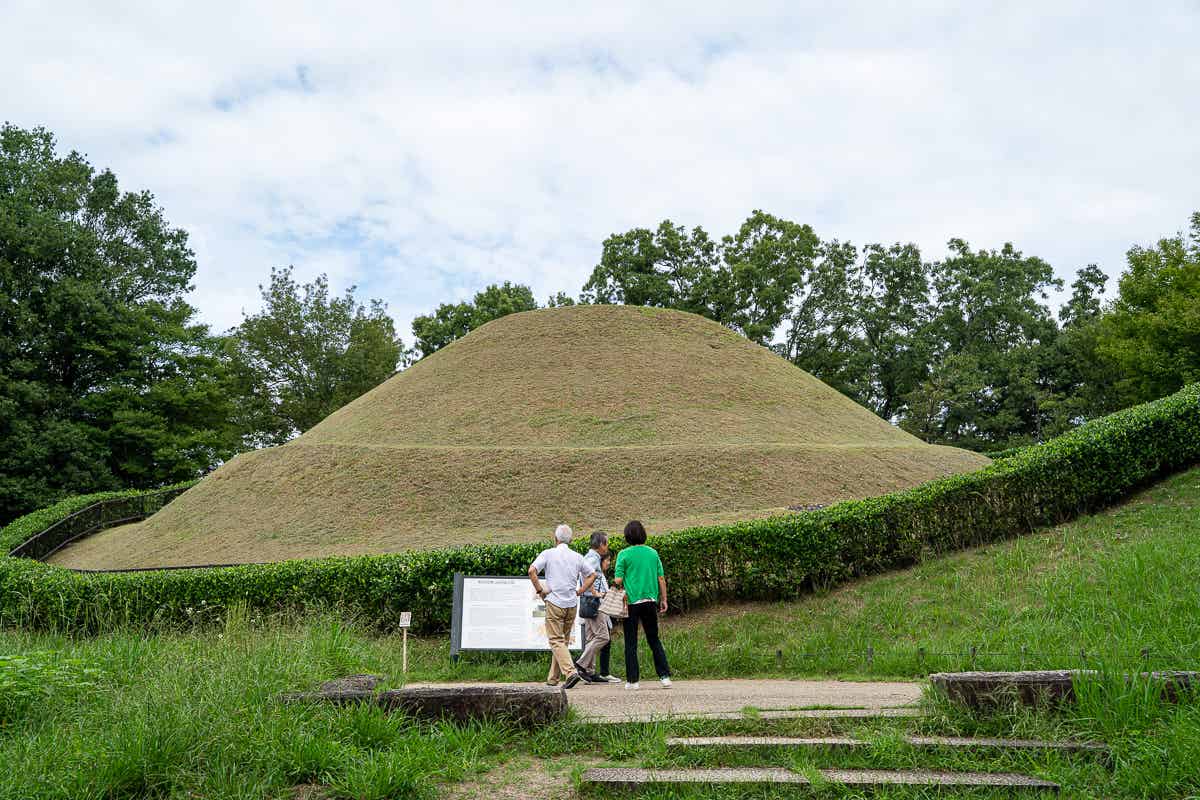 案内板の前で見学する観光客と芝生の円墳（高松塚古墳）